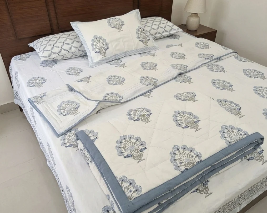 Block print bedsheet and matching folded quilt in a blue floral bouquet motif, styled with coordinating pillow shams on a bed