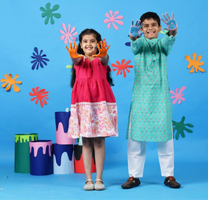 Two children in colorful traditional Indian outfits standing against a blue background with paint-splatter decorations
