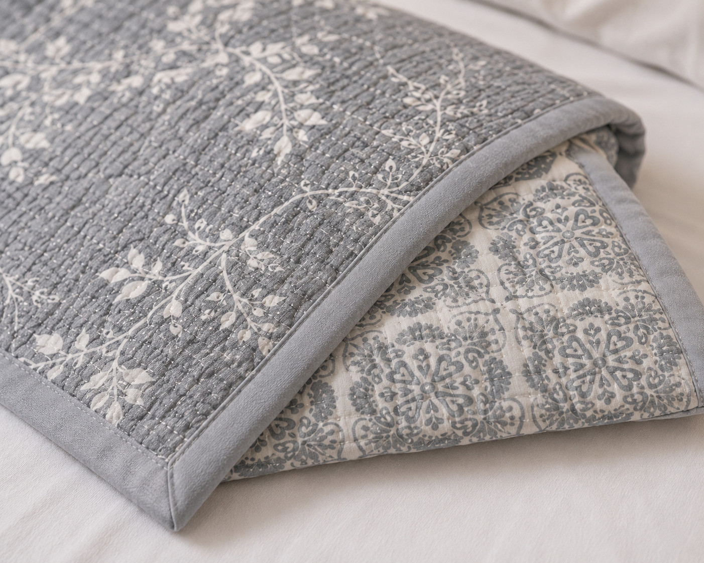 Close-up of folded grey botanical quilt showing white vine print and reverse medallion pattern with grey piping