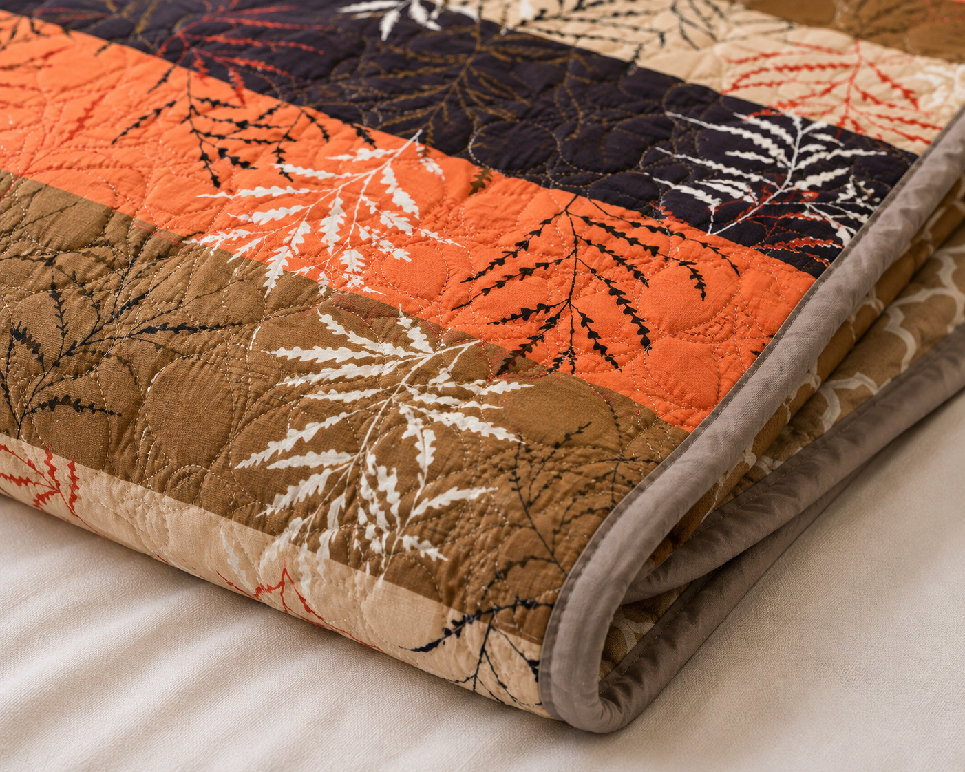 Close-up of folded single bed cotton quilted comforter showing botanical fern leaf print in orange navy and brown stripes with grey piping