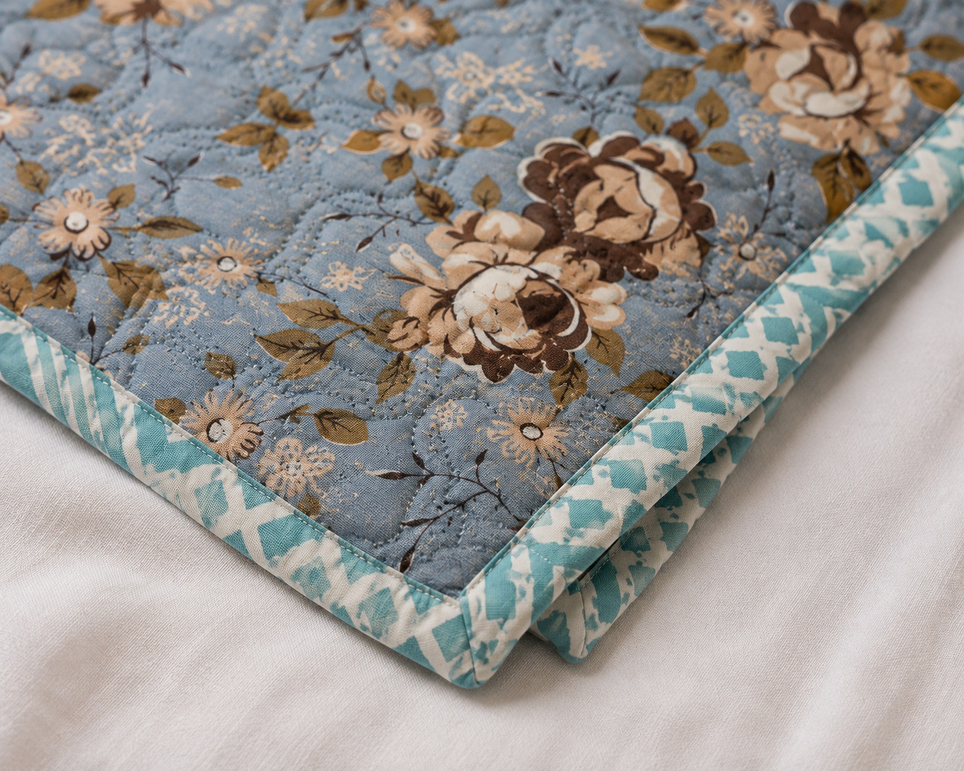 Close-up of folded single bed dusty blue floral quilted comforter showing rose print and teal diamond piping detail