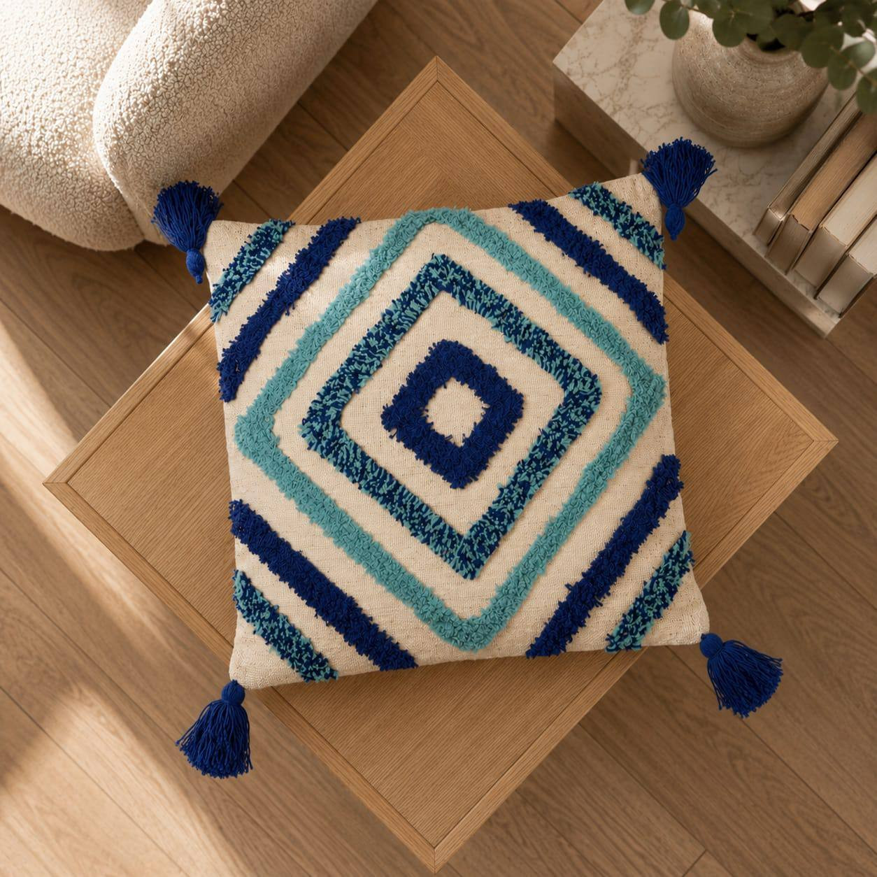 Top view of cream tufted cushion cover with blue geometric diamond design and navy tassels on wooden table
